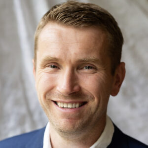 A smiling person in a blue suit and white shirt is foregrounded against a blurred gray background.