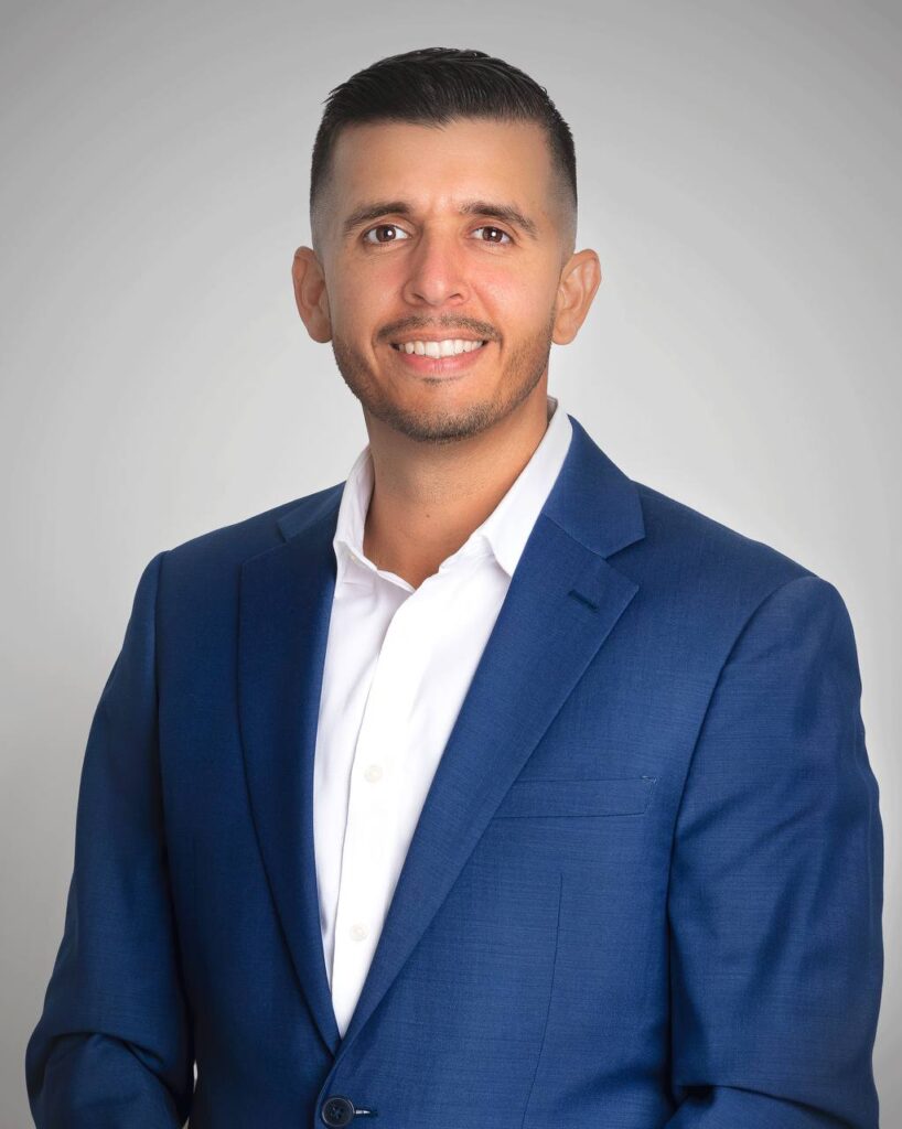 A man with short dark hair, a beard, and a bright smile is wearing a blue suit jacket over a white shirt. He is standing against a plain, light gray background.