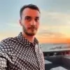 A person with short hair and a beard wearing a patterned shirt stands outdoors with a sunset in the background. The sky is a gradient of blue, orange, and pink, and a body of water is visible in the distance.